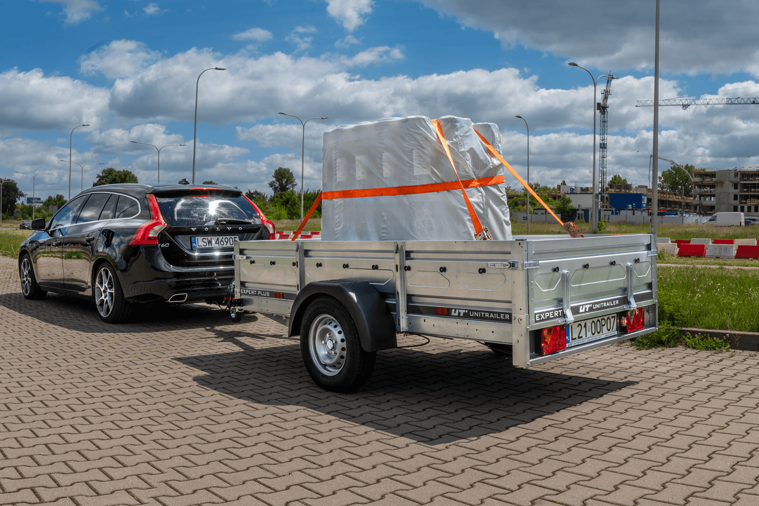 Schwarzer Volvo Kombi zieht einen einachsigen Unitrailer PKW-Anhänger, beladen mit einer weiß folierten Palette und gesichert mit orangen Spanngurten auf Pflastersteinen.