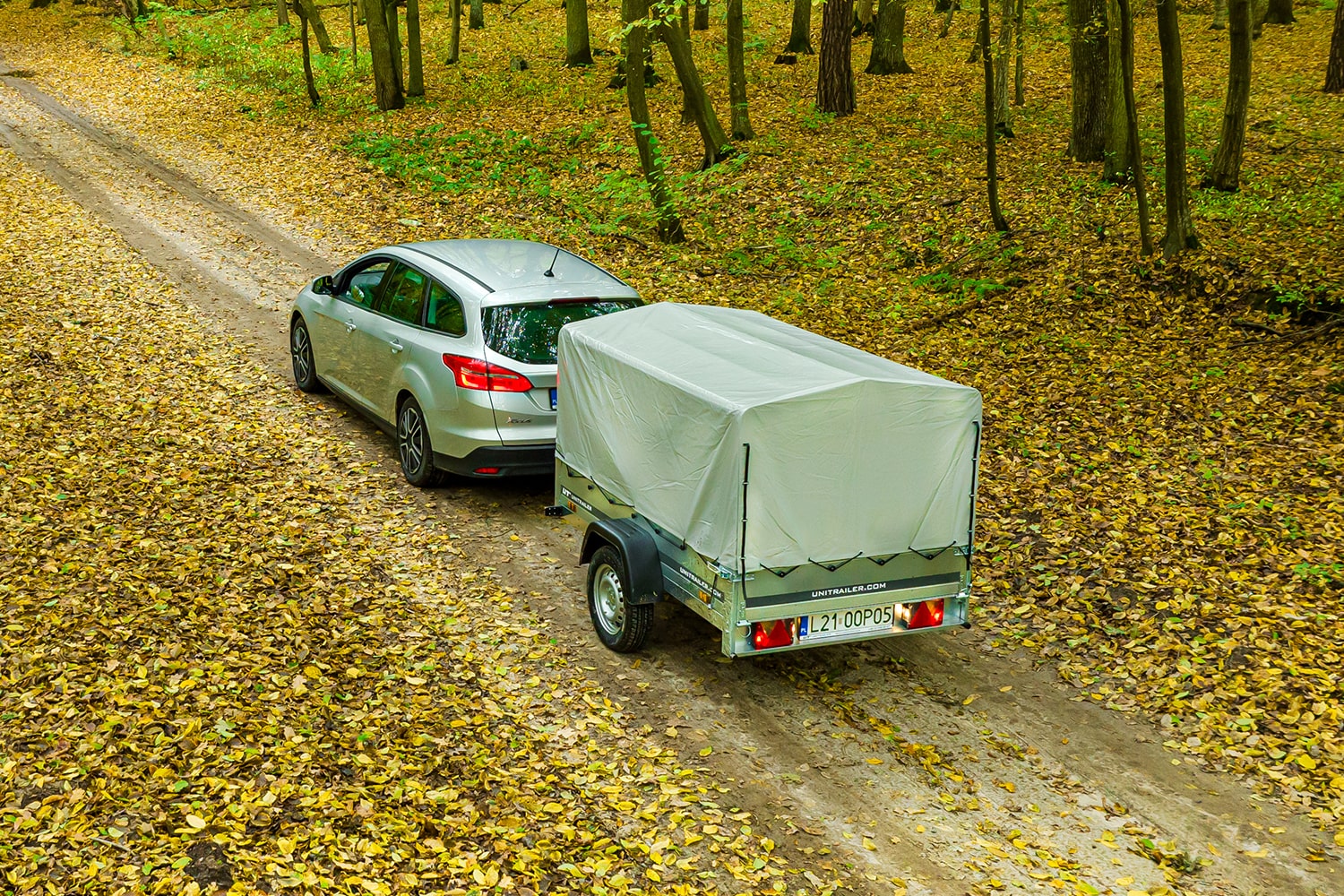 Draufsicht auf einen silbernen Kombi, der einen Unitrailer PKW-Anhänger mit hoher grauer Plane über einen mit Herbstlaub bedeckten Waldweg zieht.