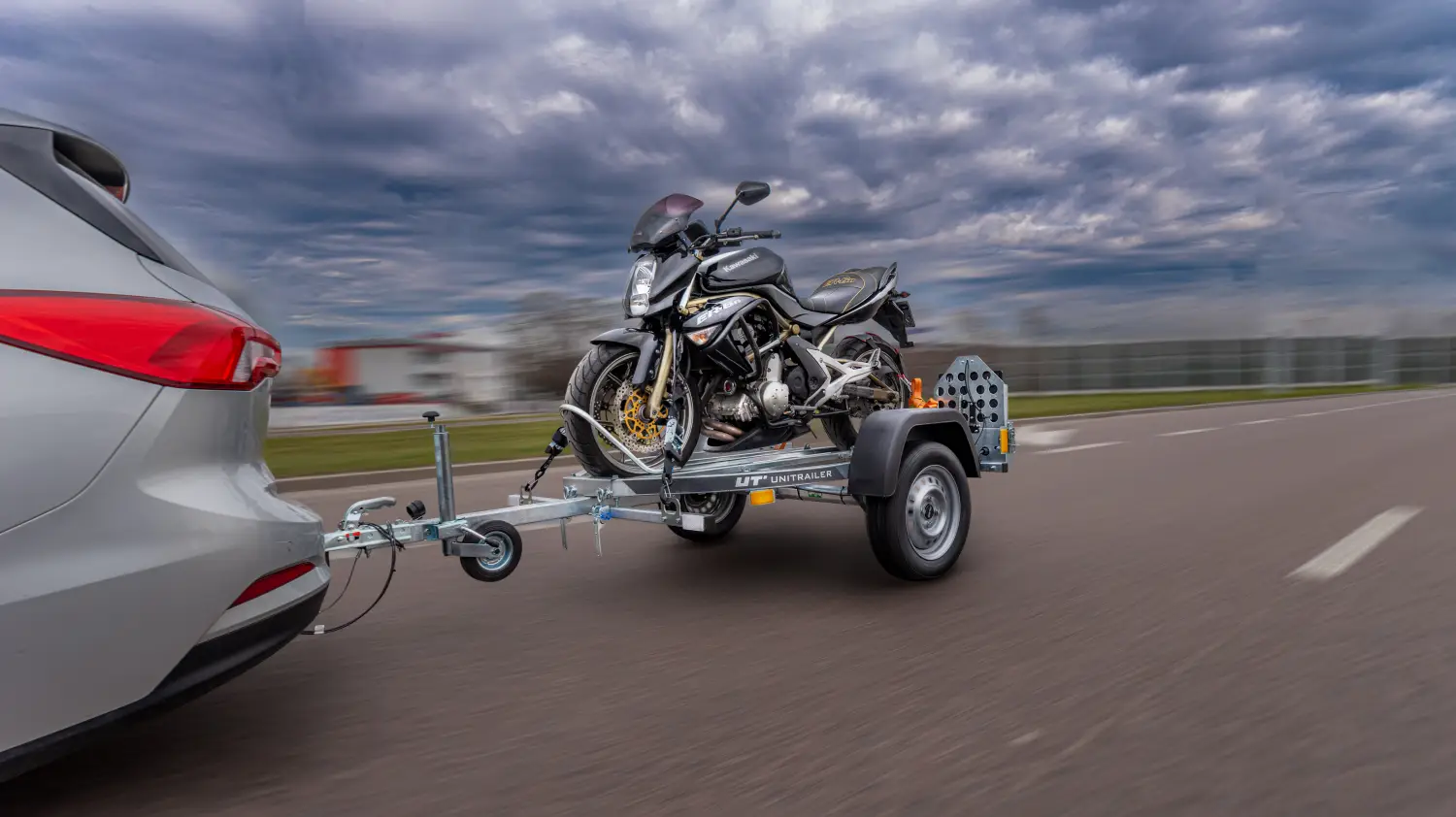 Silberner Pkw zieht einen Einachs-Motorradanhänger der Marke Unitrailer auf einer Asphaltstraße, beladen mit einem gesicherten schwarzen Kawasaki-Motorrad.