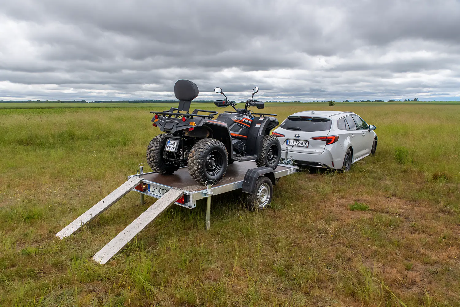 Silberner Kombi mit angehängtem Plattformanhänger auf einer grünen Wiese, beladen mit einem schwarzen Quad der Marke Goes und zwei angelegten Auffahrrampen aus Aluminium.