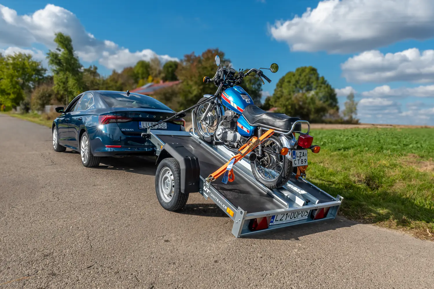 Dunkelblauer Škoda Octavia auf einer Landstraße mit einem gekippten Unitrailer-Motorradanhänger, auf dem ein klassisches blaues Honda-Motorrad mit Gurten gesichert ist.