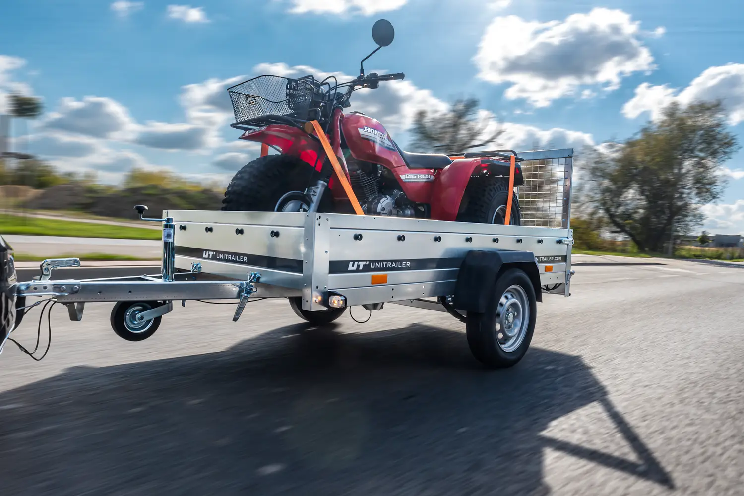 Einachsiger Kastenanhänger der Marke Unitrailer während der Fahrt, beladen mit einem roten Honda-Dreirad mit Frontkorb, gesichert durch orangefarbene Spanngurte.