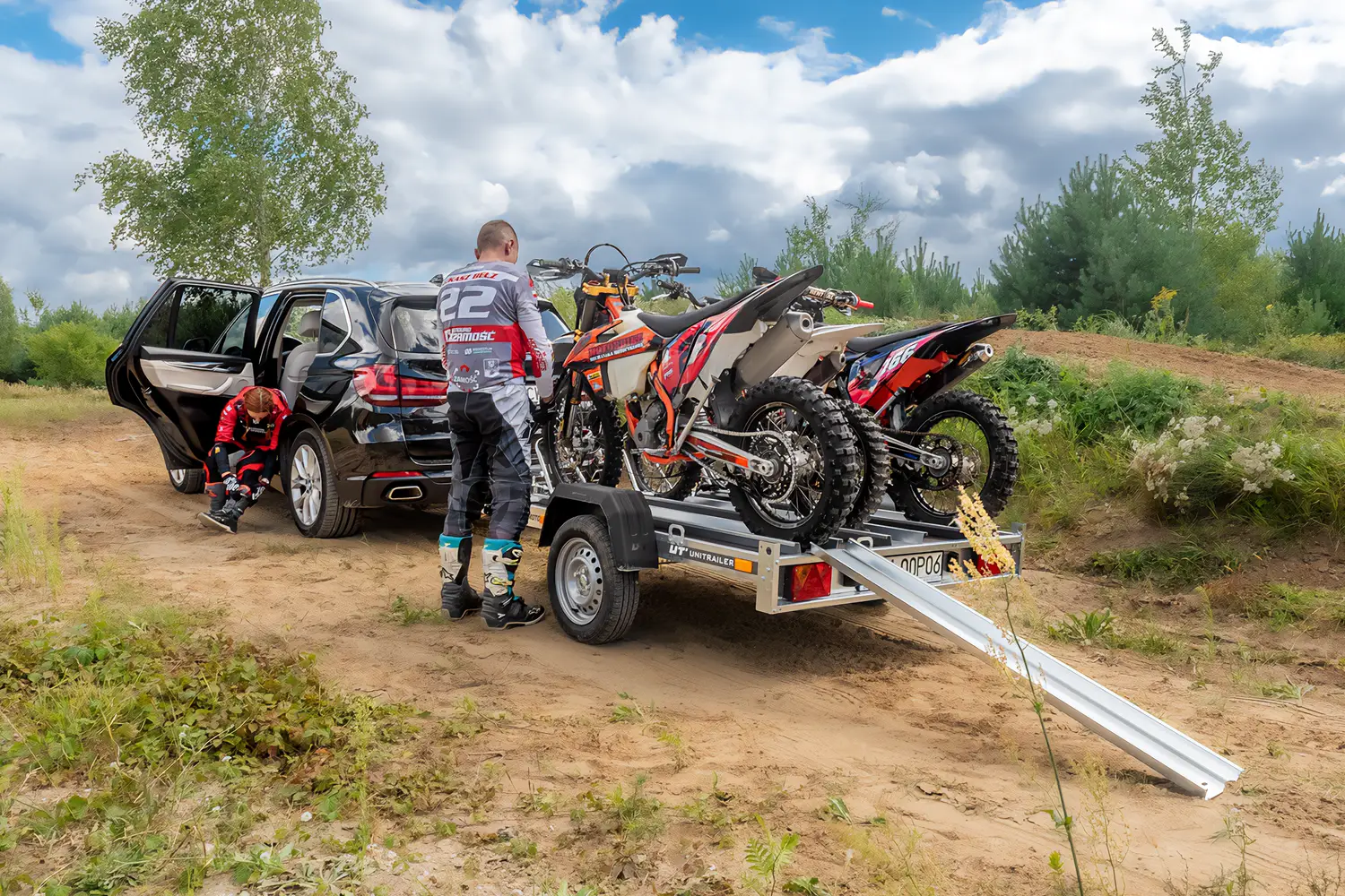 Schwarzer SUV auf einem Offroad-Gelände mit einem Unitrailer-Motorradanhänger, beladen mit zwei Motocross-Maschinen, während sich zwei Fahrer in Schutzkleidung neben der Auffahrrampe vorbereiten.