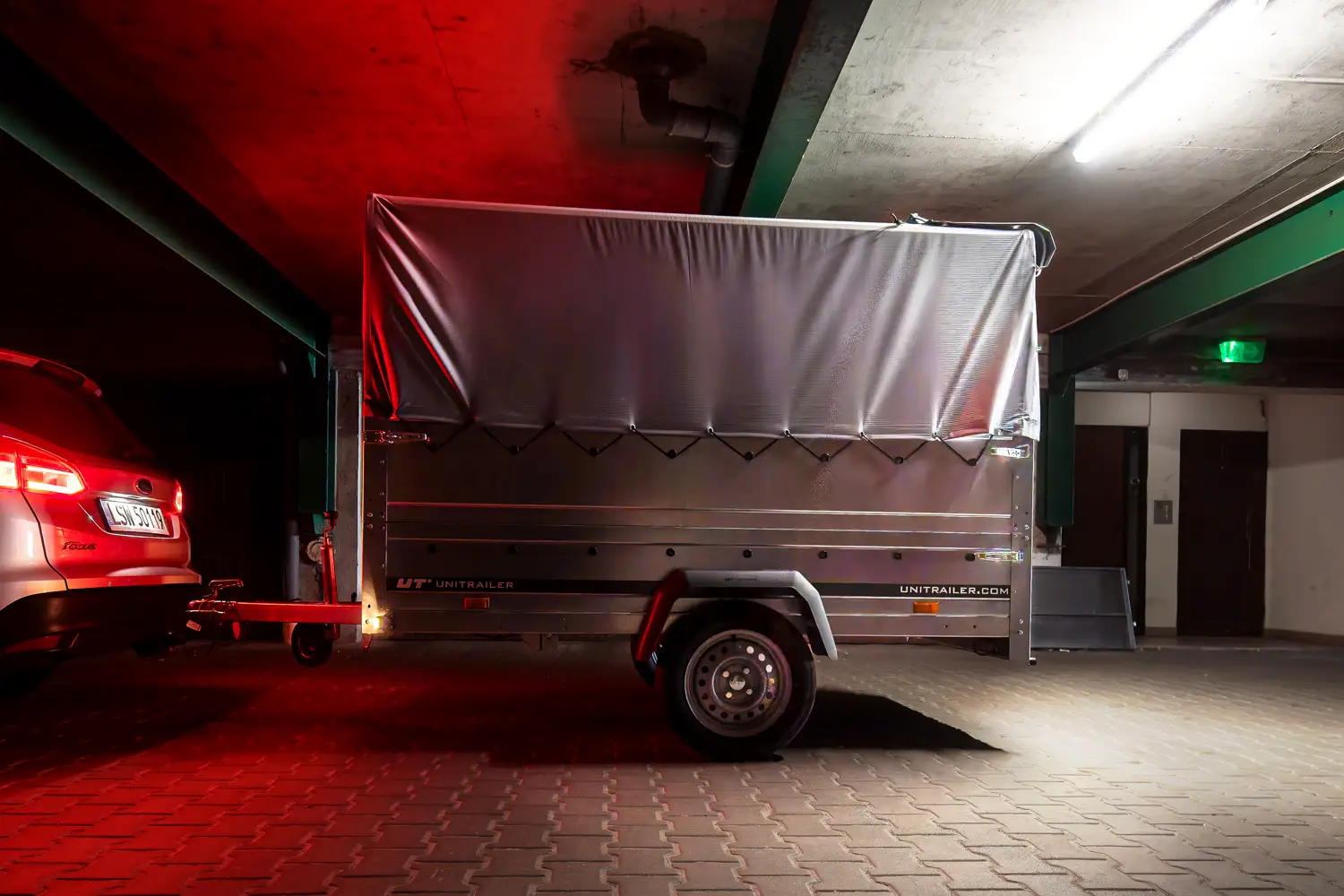 Unitrailer-Transportanhänger mit grauer Plane, an ein Auto gekuppelt, in einer schwach beleuchteten Tiefgarage.